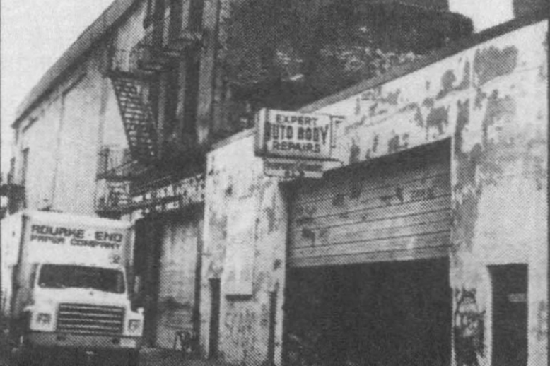 Photograph of a city block featuring a one-story white building with a garage door and a sign stating "Expert Auto Body Repairs".